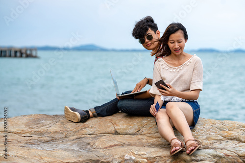 Young adult asian man and woman traveler sitting on ocean cliff on tropical island in summer day vacation holding and using laptop  smartphone and credit card for online network shopping or working.