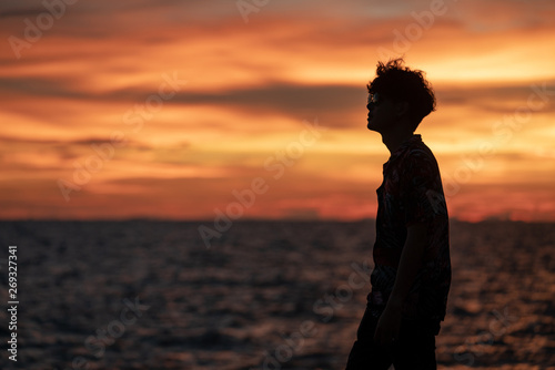 Medium long shot silhouette freedom young man traveler wearing sunglass standing on ocean island mountain cliff at summer sunset with blurred sunset twilight sky and ocean background.