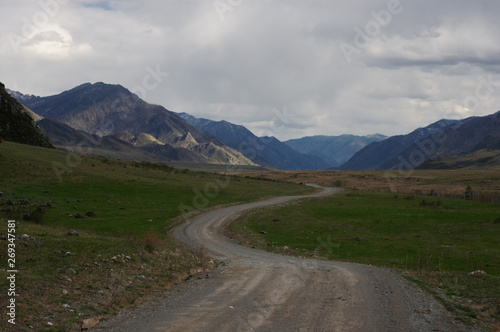 Mountain dark evening steppe valley road path with spring green grass and ranges of snow mountains rocks on a horizon skyline