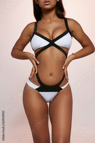 Three quarter front view shot of African lady  wearing bright white and black swimsuit with high-waisted panties. The woman is posing with hands on hips  standing against the light purple background.