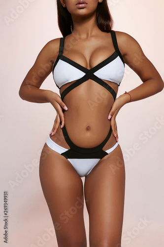 Three quarter front view shot of African lady, wearing bright white and black swimsuit with high-waisted panties. The woman is posing with hands on hips, standing against the light purple background.