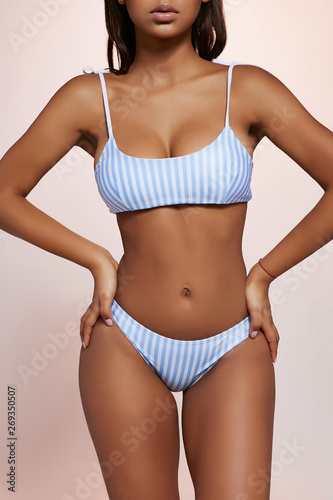 Three quarter cropped shot of African lady with straight hair, wearing stripy two-piece bikini set with scoop neckline top. The girl is standing with hands sliding down on hips on creamy background.