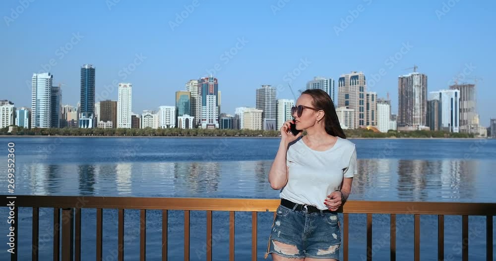 Young beautiful girl standing on the waterfront against the city talking on the phone Young beautiful girl standing on the waterfront against the city talking on the phone
