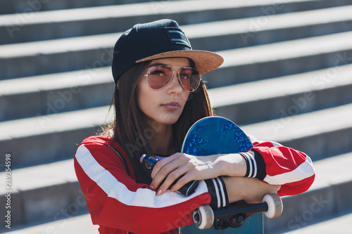 Vaping girl. Young woman with skateboard vape e-cig. Pretty young female in black hat, red clothing vape ecig, vaping device at the sunset. Toned image. Hip-hop style. photo