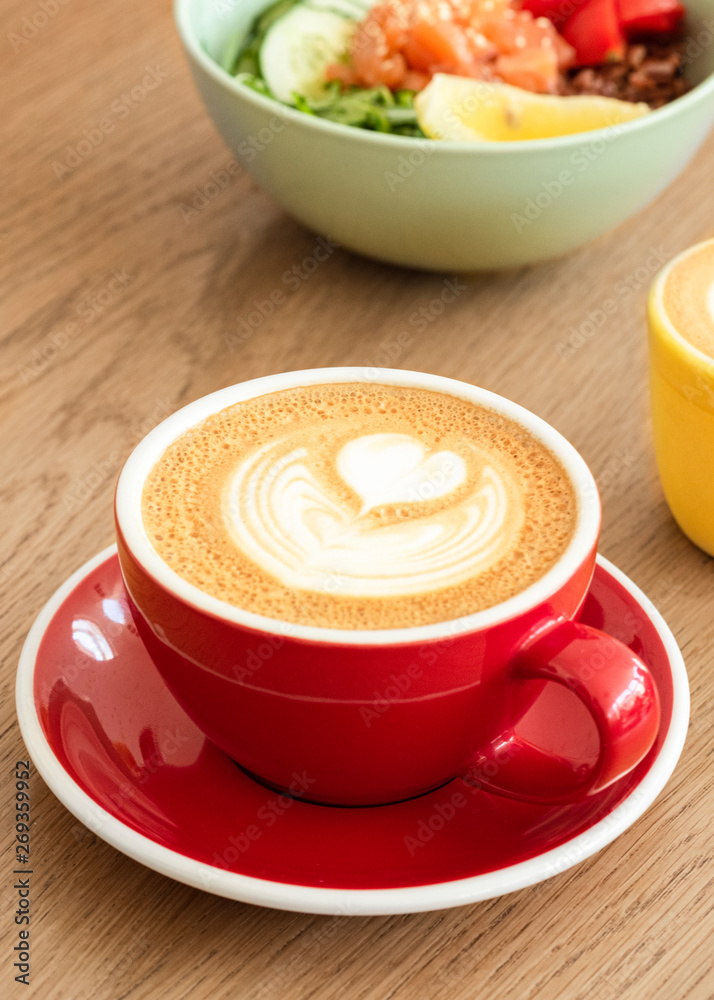 A red cup of cappuccino with trendy latte art on a saucer with a coffee spoon. In the background a healthy breakfast bowl. A red cup of cappuccino with trendy latte art on a saucer with a coffee spoon. In the background a healthy breakfast bowl.