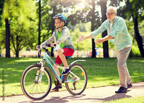 family, generation, safety and people concept - happy grandfather teaching boy how to ride bicycle at summer park