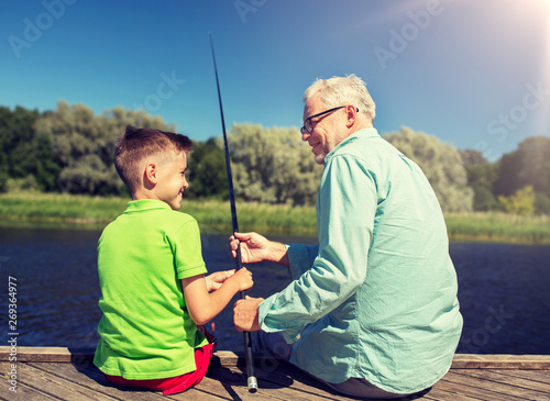 family, generation, summer holidays and people concept - happy grandfather and grandson with fishing rods on river berth