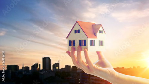 real estate, accommodation and property concept - close up of hand holding house or home model over sunset sky in tallinn city, estonia background