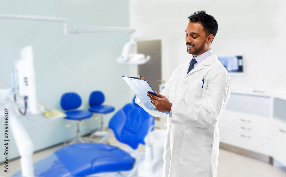 medicine, dentistry and profession concept - smiling indian male dentist in  white coat with clipboard over dental clinic office background Stock Photo  | Adobe Stock