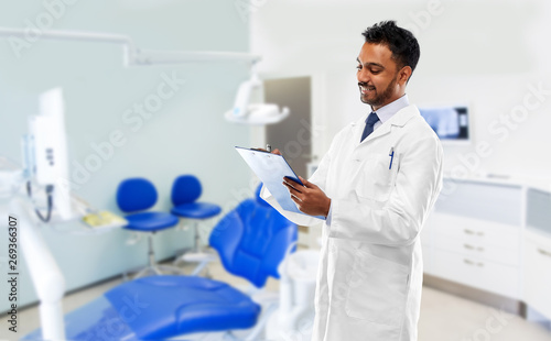 medicine  dentistry and profession concept - smiling indian male dentist in white coat with clipboard over dental clinic office background
