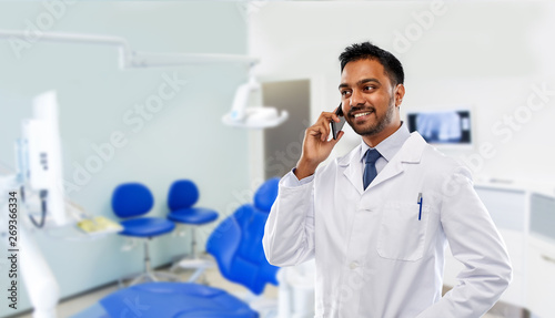 medicine, dentistry and healthcare concept - smiling indian male dentist in white coat calling on smartphone over dental clinic office background