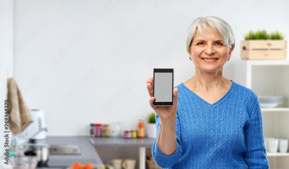 technology and old people concept - smiling senior woman showing smartphone over kitchen background technology and old people concept - smiling senior woman showing smartphone over kitchen background