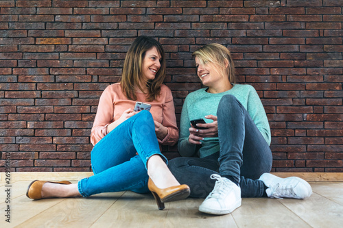 two beautiful smiling women sitting on the floor looking to each other holding a mobile phone on their hands