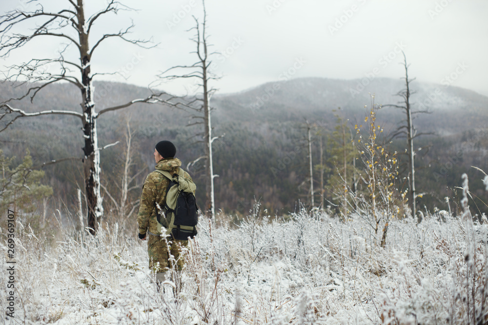 Man in camouflage with a backpack is standing on a snow-covered mountainside among bushes and trees. Extreme travel. Survival in the wild. Man in camouflage with a backpack is standing on a snow-covered mountainside among bushes and trees. Extreme travel. Survival in the wild.