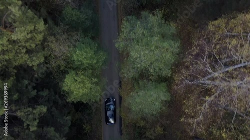 Aerial; drone footage of black car moving on the road through the autumn forest; view from above of green pine and yellow birch trees crones; cloudy sunset; autumn evening in Ural; travelling
