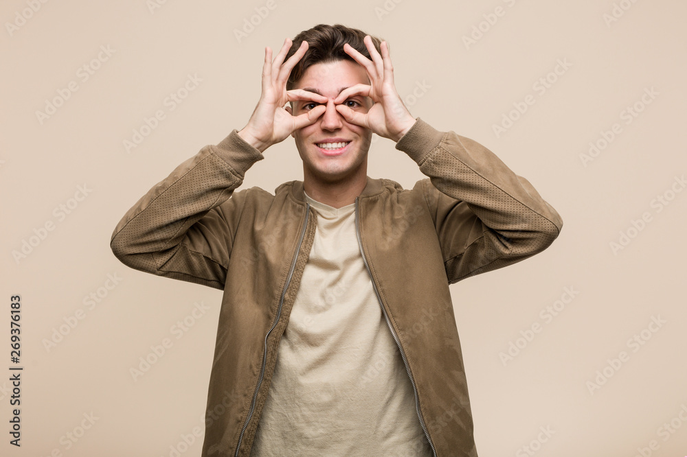 Young caucasian man wearing a brown jacket showing okay sign over eyes Young caucasian man wearing a brown jacket showing okay sign over eyes