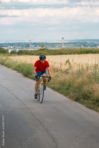 A cyclist in red blue form rides on a road bicycle along field.