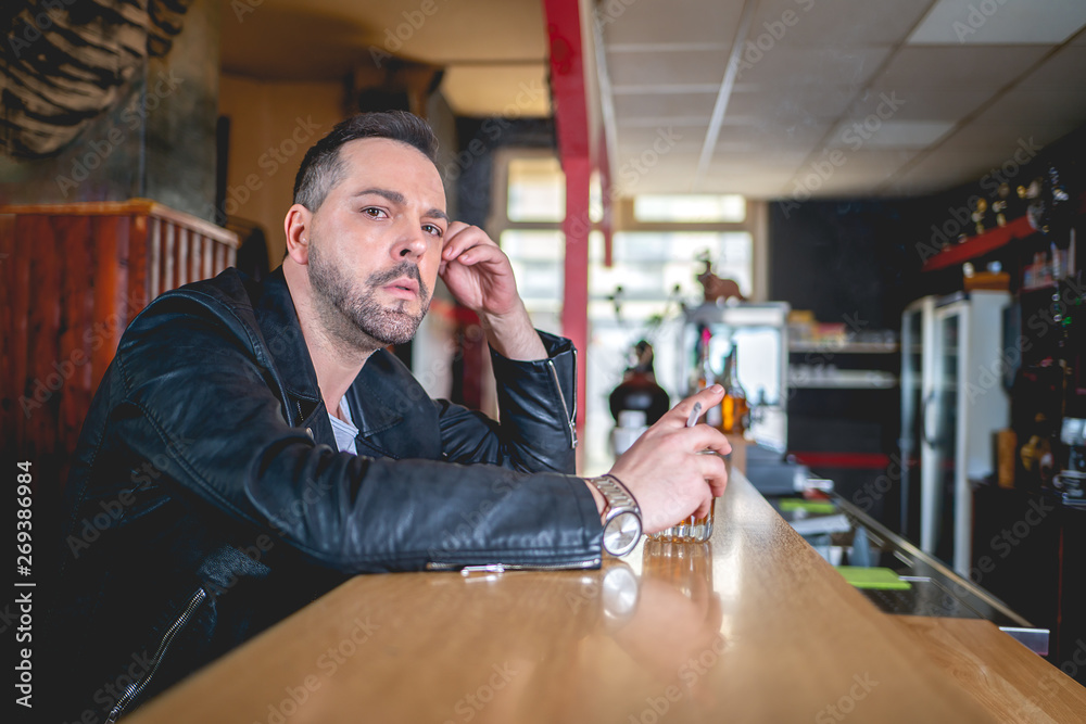 A man with a drink and a cigarette at a bar looks thoughtfully at the camera