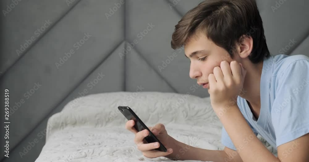 Teenager typing text message by smart phone, at home. Handsome boy spending time in a social network using mobile phone lying on sofa. Caucasian teenage guy talking on cell phone. Lifestyle concept.