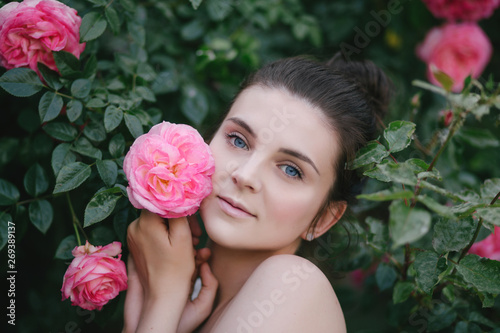Young beautiful woman portrait close up with perfect skin posing with pink roses flowers in a garden © polinaloves