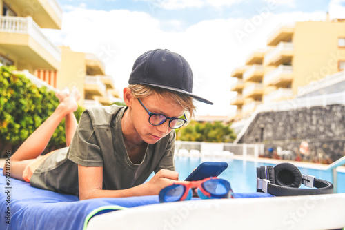 Young boy relaxing outdoor playing with smartphone. Trendy child reads news near the swiming pool on a sunny summer day. Teen enjoying holidays after the end of school. Concept of leisure and freetime