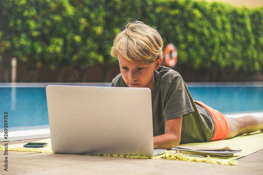 Beautiful young boy using laptop near the swimming pool in a sunny summer day Child doing homeworks outdoors after the end of school. Technology allows to stay connected with remote friends everywhere