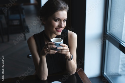Portrait of gorgeous female drinking tea or coffee and looking with smile out of the coffee shop window while enjoying her leisure time, nice business woman lunch in modern cafe during her work break.