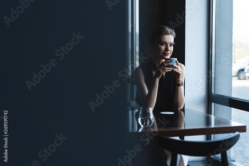 Portrait of gorgeous female drinking tea or coffee and looking with smile out of the coffee shop window while enjoying her leisure time, nice business woman lunch in modern cafe during her work break.