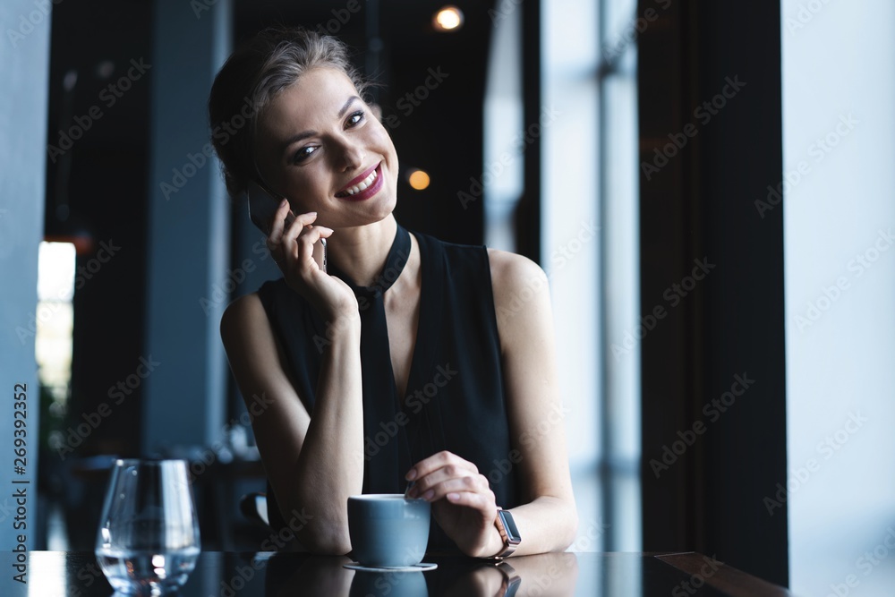 Fototapeta premium Young charming woman calling with cell telephone while sitting in coffee shop during free time, attractive female having talking conversation with mobile phone while rest in cafe.