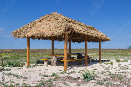 Wooden gazebo with a roof of reeds in the steppe. On the bench is a tourist backpack. Blue sky  steppe grass and flowers.