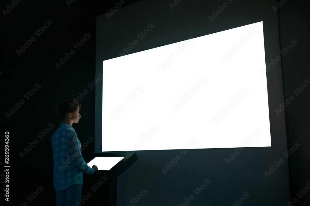Woman using electronic kiosk and looking at white blank large interactive wall display in dark room of modern technology exhibition. Mock up, futuristic, template, education and technology concept Woman using electronic kiosk and looking at white blank large interactive wall display in dark room of modern technology exhibition. Mock up, futuristic, template, education and technology concept