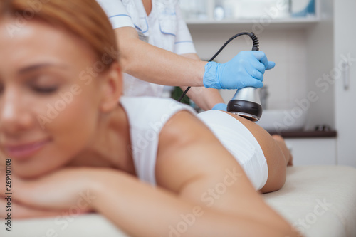 Selective focus on ultrasound cavitation machine professional cosmetologist is using on female client. Woman receiving anti-cellulite slimming procedure at beauty salon photo