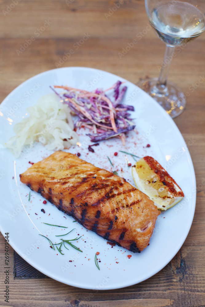 Delicious grilled salmon fish steak served with seasonings, veggies and lemon on a white plate on a wooden table. Restaurant menu design. Barbecue party. 