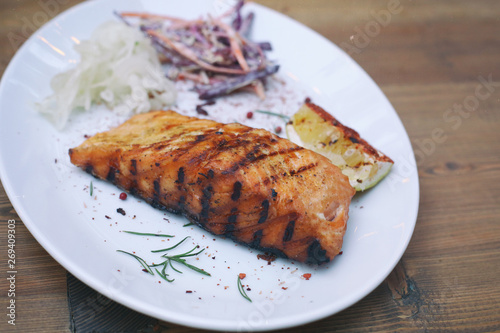 Delicious grilled salmon fish steak served with seasonings, veggies and lemon on a white plate on a wooden table. Restaurant menu design. Barbecue party. 