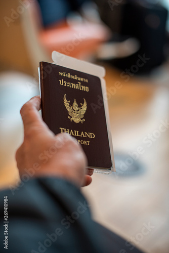 Male holding a Thailand passport with a boarding pass attached. He checks his passport while sitting in a front row waiting for boarding time near the air port gate during morning time.