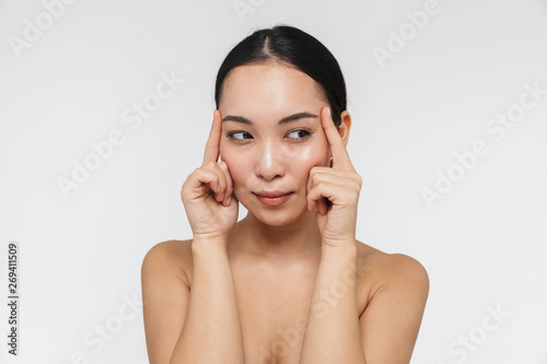 Portrait of smiling half naked asian woman touching her clear skin and looking aside