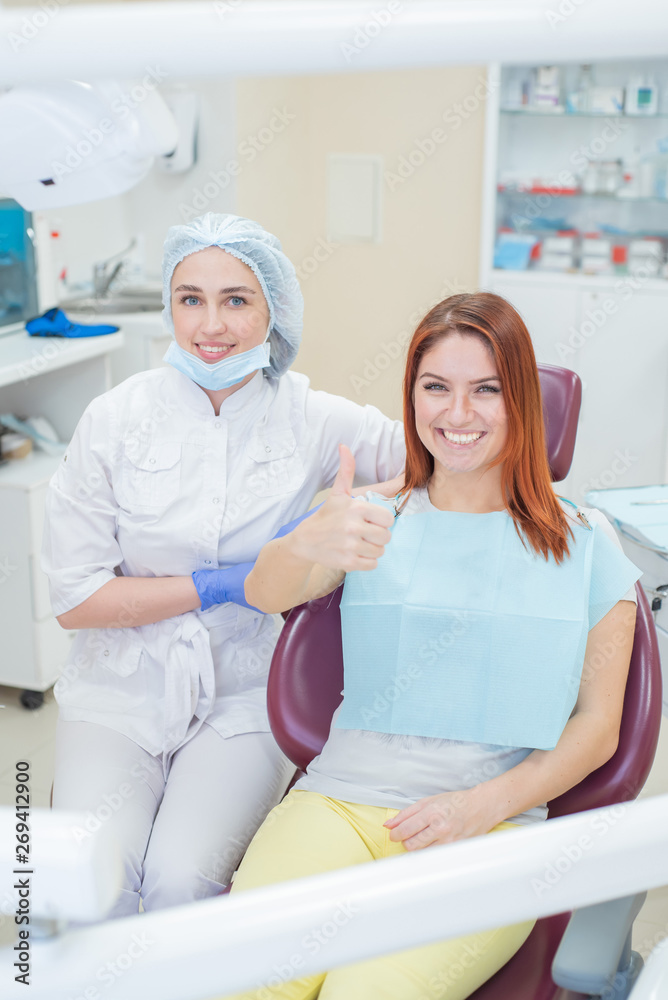 Obraz premium Happy women a dentist and patient after treating teeth at the dental office, smiling and looking towards the camera. Happy patient shows thumb as an endorsement. Dental equipment.