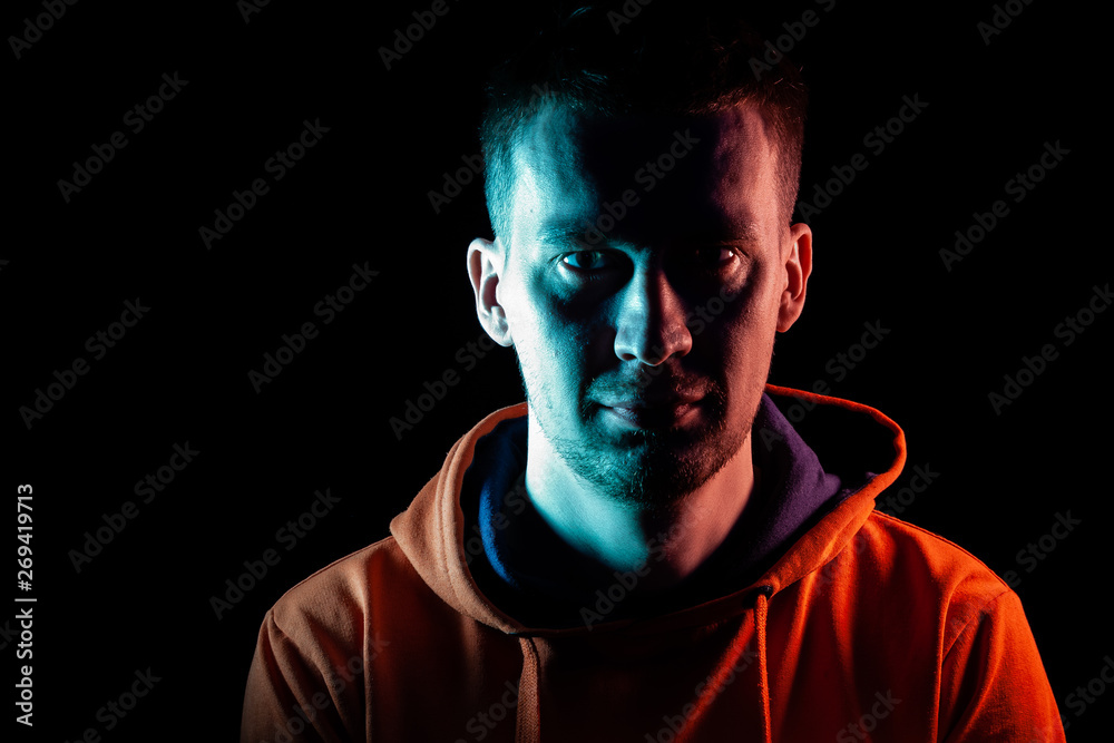 An adult guy stands look forward in an orange hooded sweatshirt highlighted in blue and pink on the sides with a smile on a black isolated background.