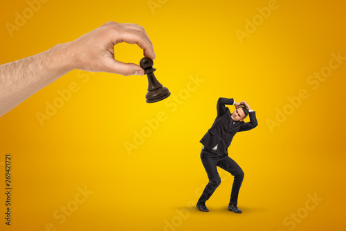 Little scared businessman covering his head with hands to protect from huge hand holding black chess pawn on yellow background.