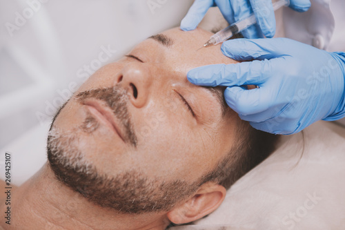 Close up of a handsome mature bearded man receiving face injections at beauty clinic. Professional cosmetologist doing wrinkle filler injections for male client