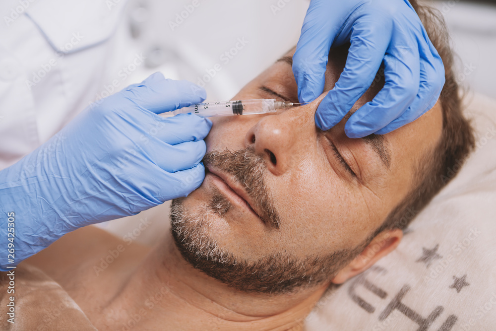 Mature man getting liquid rhinoplasty by professional cosmetologist. Attractive male client getting non-surgical nose job at cosmetology clinic. Beautician injecting filler in the face of mature man Mature man getting liquid rhinoplasty by professional cosmetologist. Attractive male client getting non-surgical nose job at cosmetology clinic. Beautician injecting filler in the face of mature man