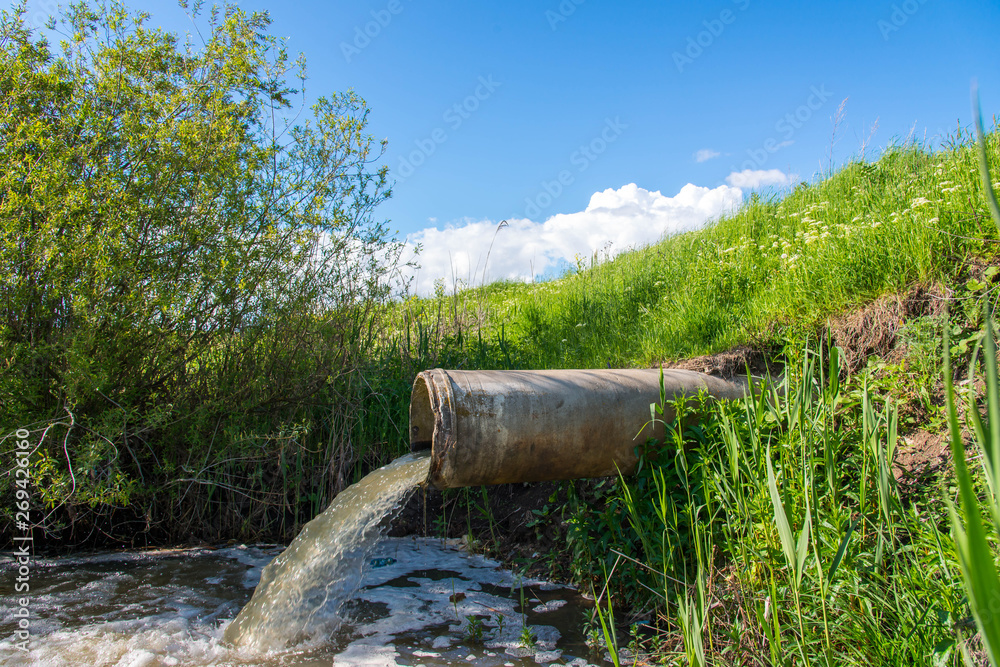 Concrete pipe transporting the polluted sewage water in to a small pond, blue sky with white clouds in the background.