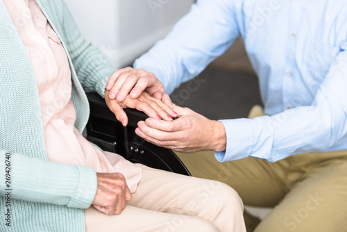 partial view of man holding hands with disabled senior mother on wheelchair