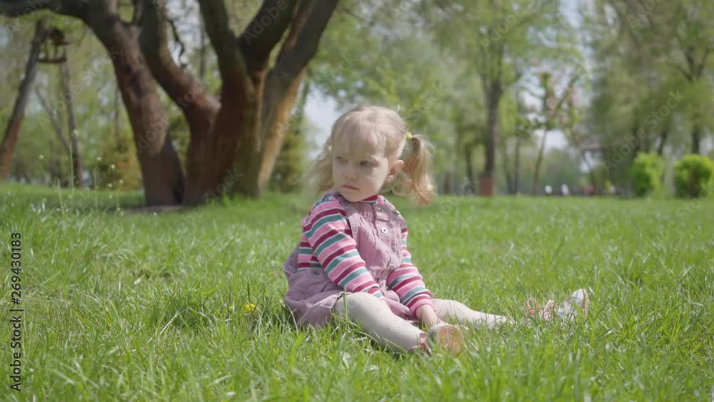Cute little girl sitting on the grass in the park, playing alone, pointing with a tiny finger up. Carefree child resting outdoors. Active healthy lifestyle, connection with nature Cute little girl sitting on the grass in the park, playing alone, pointing with a tiny finger up. Carefree child resting outdoors. Active healthy lifestyle, connection with nature