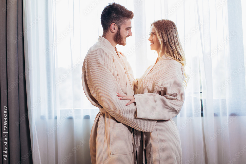 Young happy couple in hotel room in the morning. Just married man and woman standing at window. Young happy couple in hotel room in the morning. Just married man and woman standing at window.