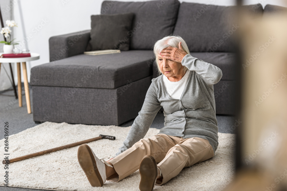 senior woman with migraine sitting on carpet and touching forehead with hand