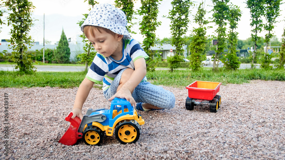 Closeup portrait of happy smiling 3 years old child boy digging sand on the playground with toy plastic truck or excavator. Child palying and having fun at park at summer Closeup portrait of happy smiling 3 years old child boy digging sand on the playground with toy plastic truck or excavator. Child palying and having fun at park at summer