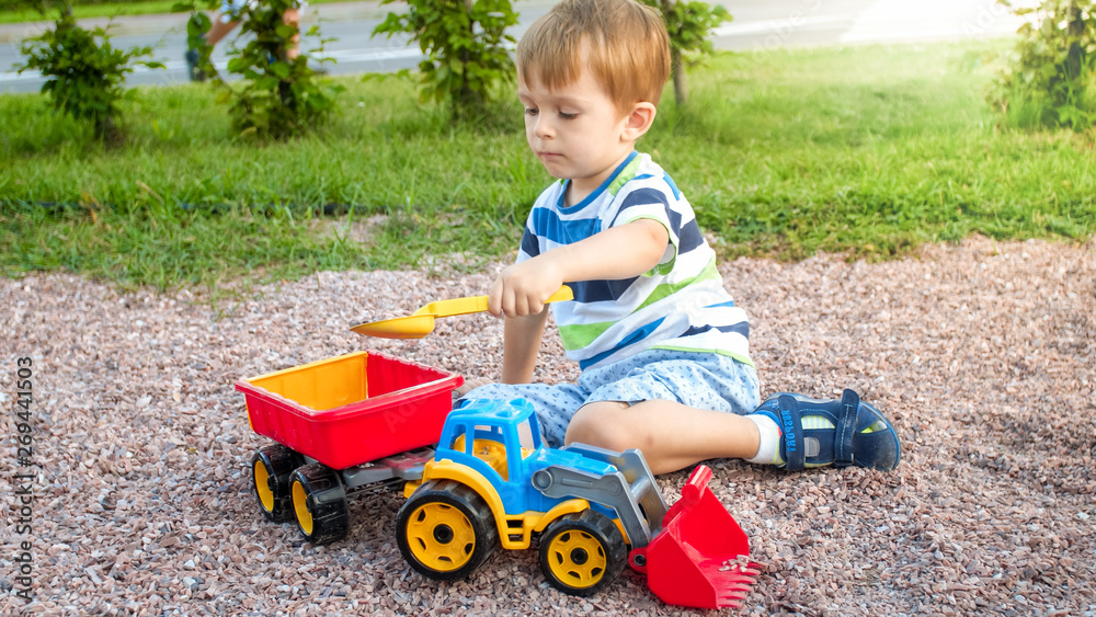 Closeup image of cute little boy playing on the palyground with toys. Child having fun with truck, excavator and trailer. He is pretending to be a builder or driver Closeup image of cute little boy playing on the palyground with toys. Child having fun with truck, excavator and trailer. He is pretending to be a builder or driver