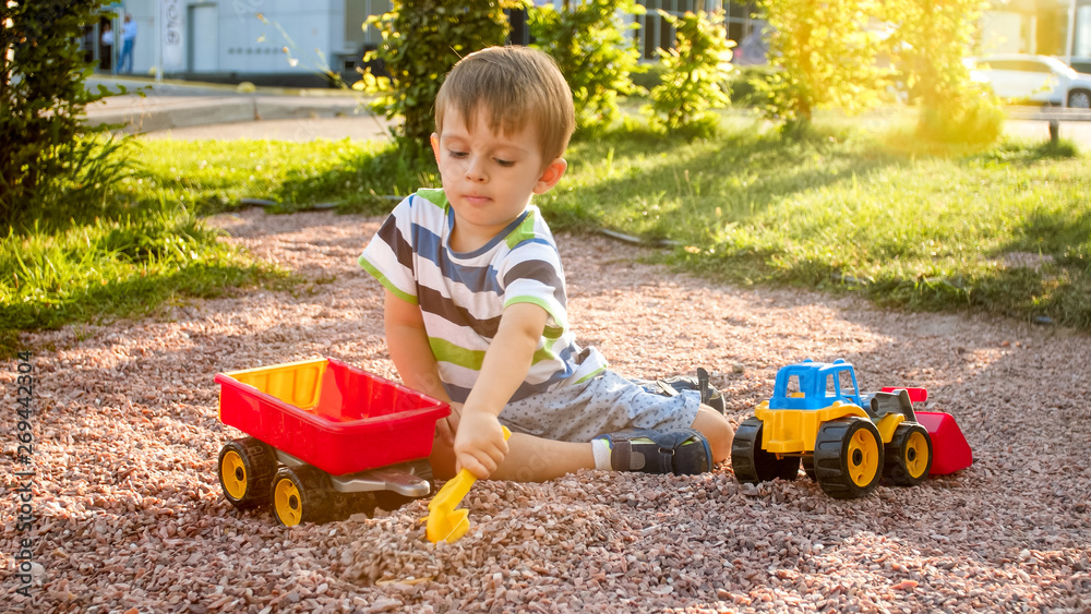 Photo of adorable 3 years old toddler boy playing with sand and you truck and trailer in park. Child digging and building in sandpit