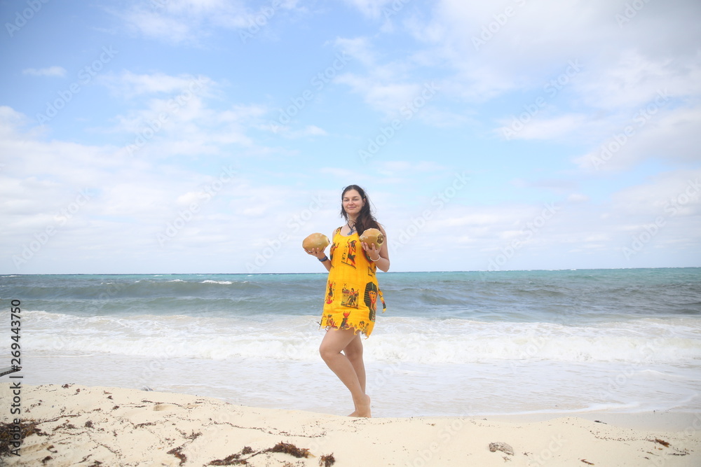 Summer holidays and vacations - an attractive girl on the sandy beach of the sea with coconuts in her hands in a yellow dress against a blue sea and sky